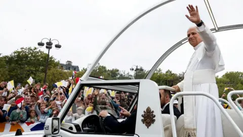 Reuters Pope Leo smiles and holds his arm up in his popemobile. Crowds can be seen waving flags and gesturing towards the pope. Some are holding up their phones or cameras to take pictures