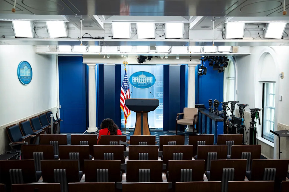 The White House Briefing Room is empty except for one reporter sitting in the front row wearing a red blazer and facing away from the camera.