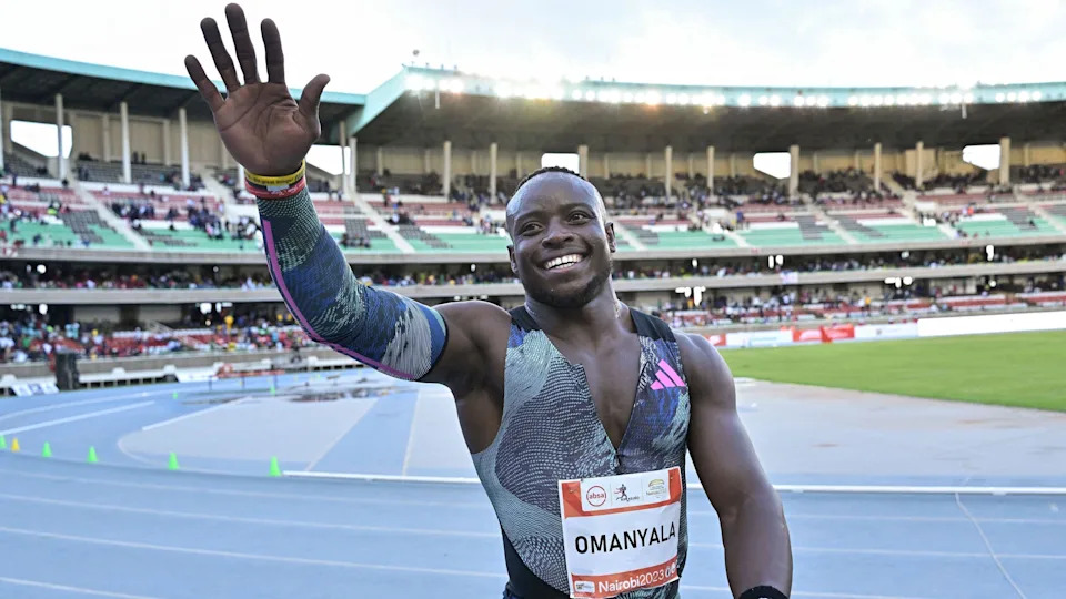 Ferdinand Omanyala, visible from waist up wearing matching athletics vest, unzipped over the chest, and sleeve over his right arm, smiles and waves to the crowd with his right arm raised while standing on the blue running track at the Kasarani Stadium. Sparsely populated stands can be seen running around the bend of the track behind him, with floodlights twinkling despite the fact that the sky is not yet dark