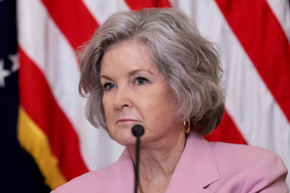 Susie Wiles wearing a pink blazer speaking at a microphone, with blurred U.S. flags in the background.
