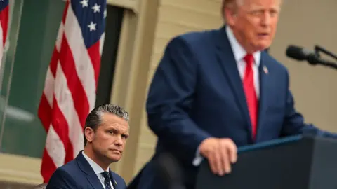 Getty Images Pete Hegseth seated while President Trump stands at the lectern in a blue suit and red tie