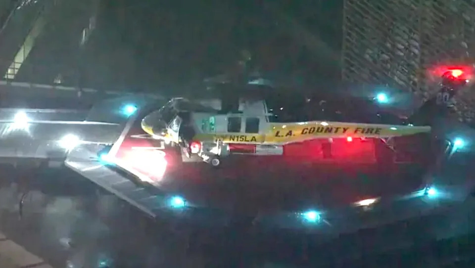 A helicopter with the words 'L.A. County Fire' at night, flying above an area marked with blue lights